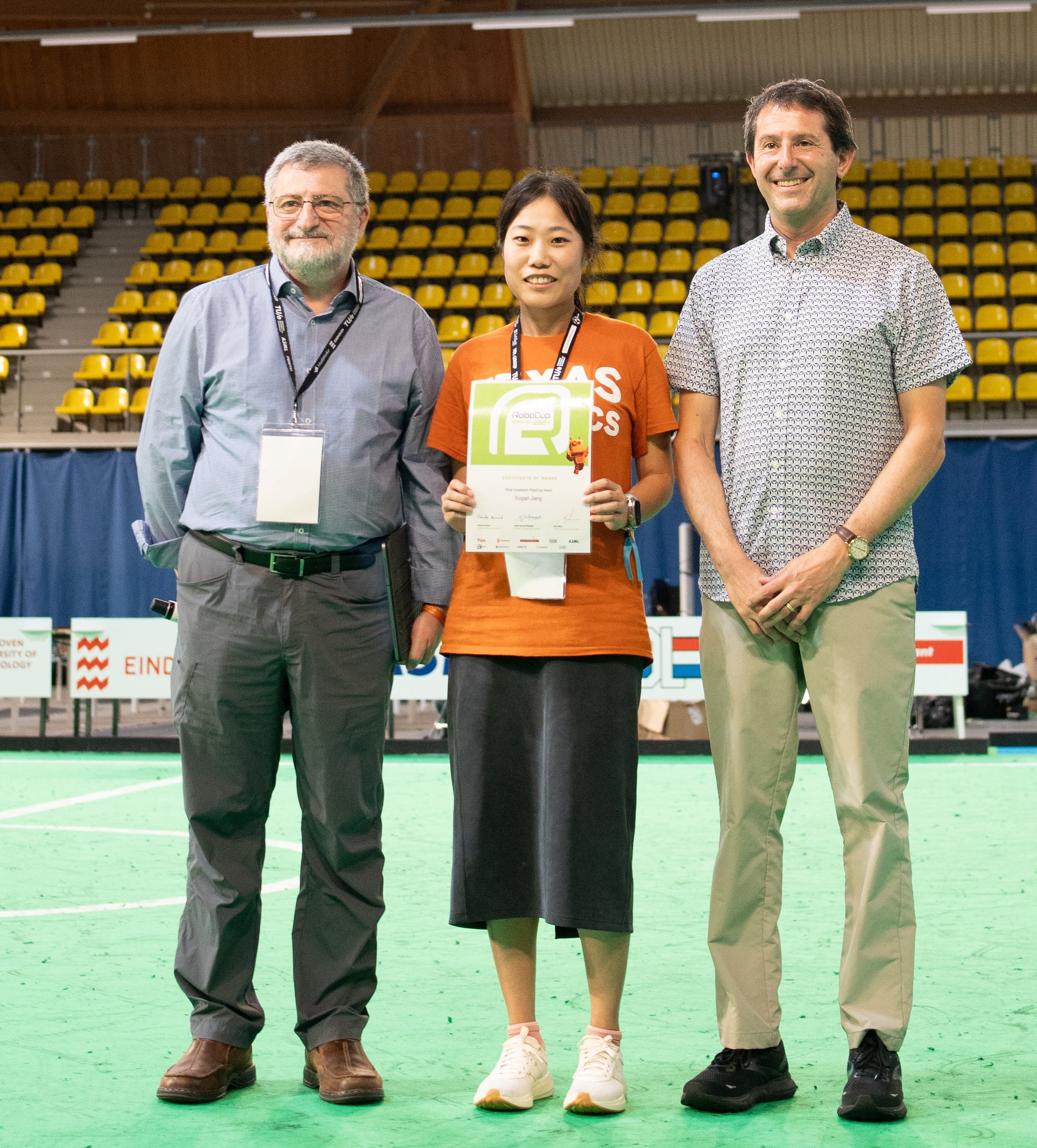 A woman in a Texas Robotics shirt holds up a certificate while posing between two men on an indoor soccer field.