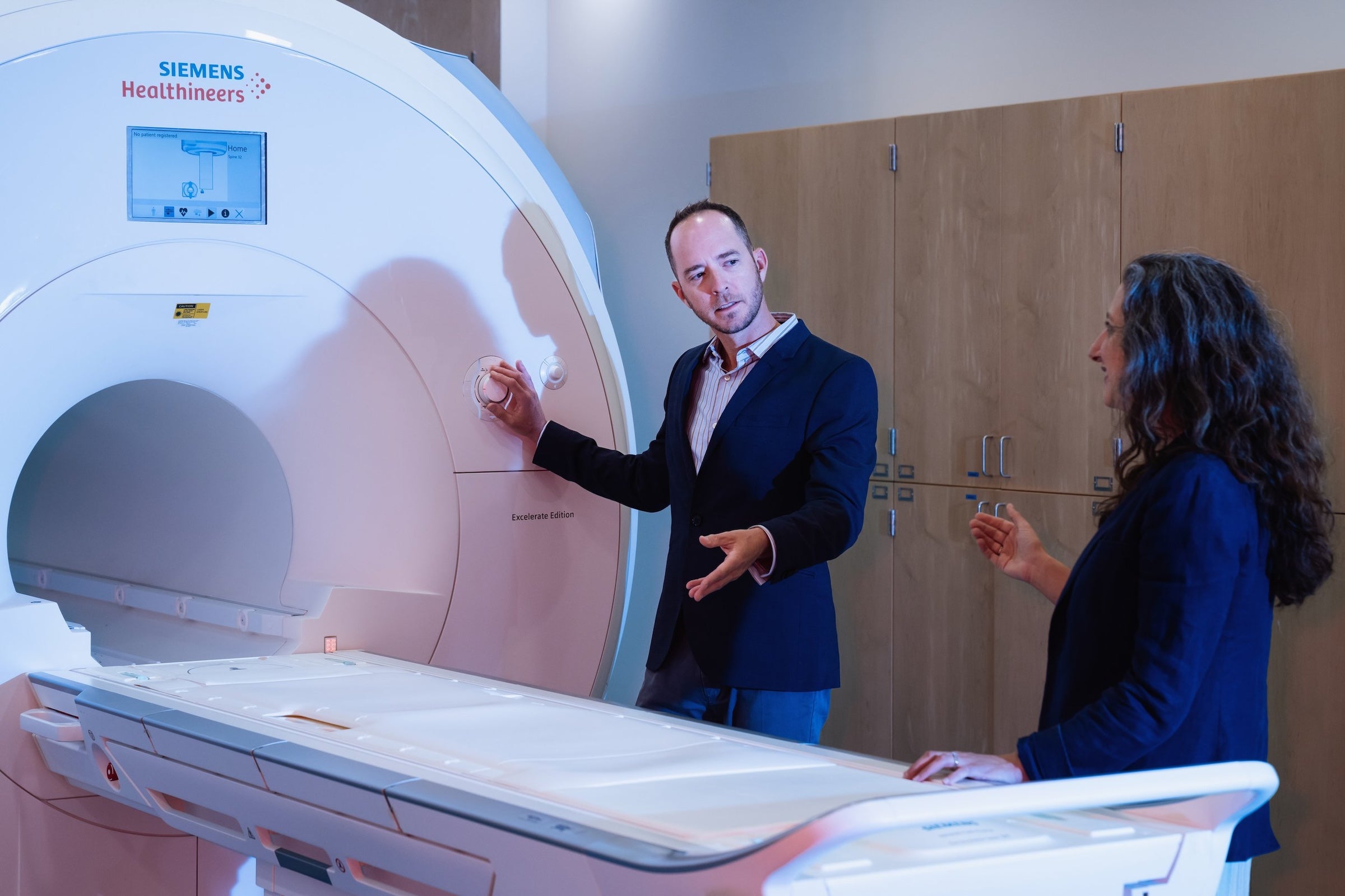 A man and woman, each wearing blazers gesture with their hands as they converse near an MRI machine